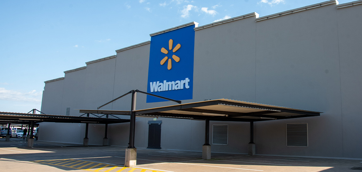 A large Walmart retail store exterior is shown under a clear blue sky. The building features a prominent blue sign with the Walmart name and icon mounted high on a plain gray facade. A covered drive-up or loading area extends from the front of the structure, casting shadows on the pavement. Yellow and white markings are visible on the ground near the entrance, suggesting designated traffic or parking zones.