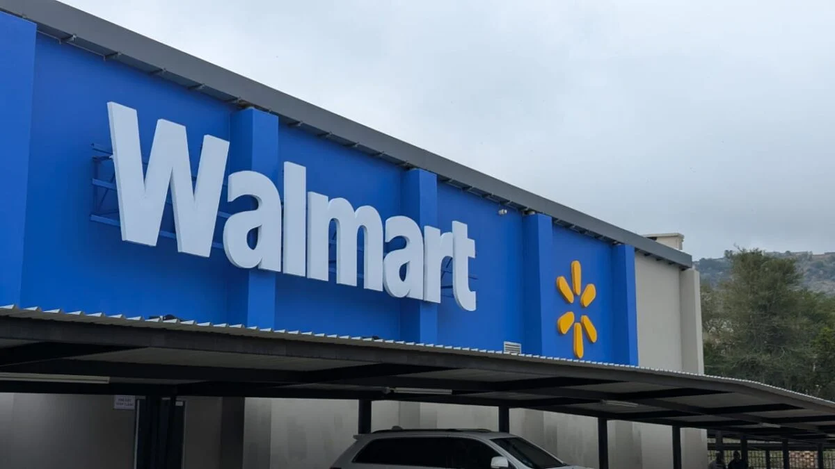 A Walmart storefront featuring a large blue sign with the brand name and logo prominently displayed. The setting includes a parking area with a visible vehicle and a cloudy sky.
