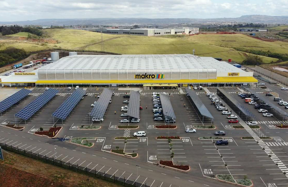A large Makro wholesale store is shown with a spacious parking lot covered by solar panel structures. The setting is an open, semi-rural area with rolling hills in the background. Numerous cars are parked under the solar panels, and clear signage such as 'STOP' is visible on the pavement. The overall mood is utilitarian and modern, with a focus on sustainability.