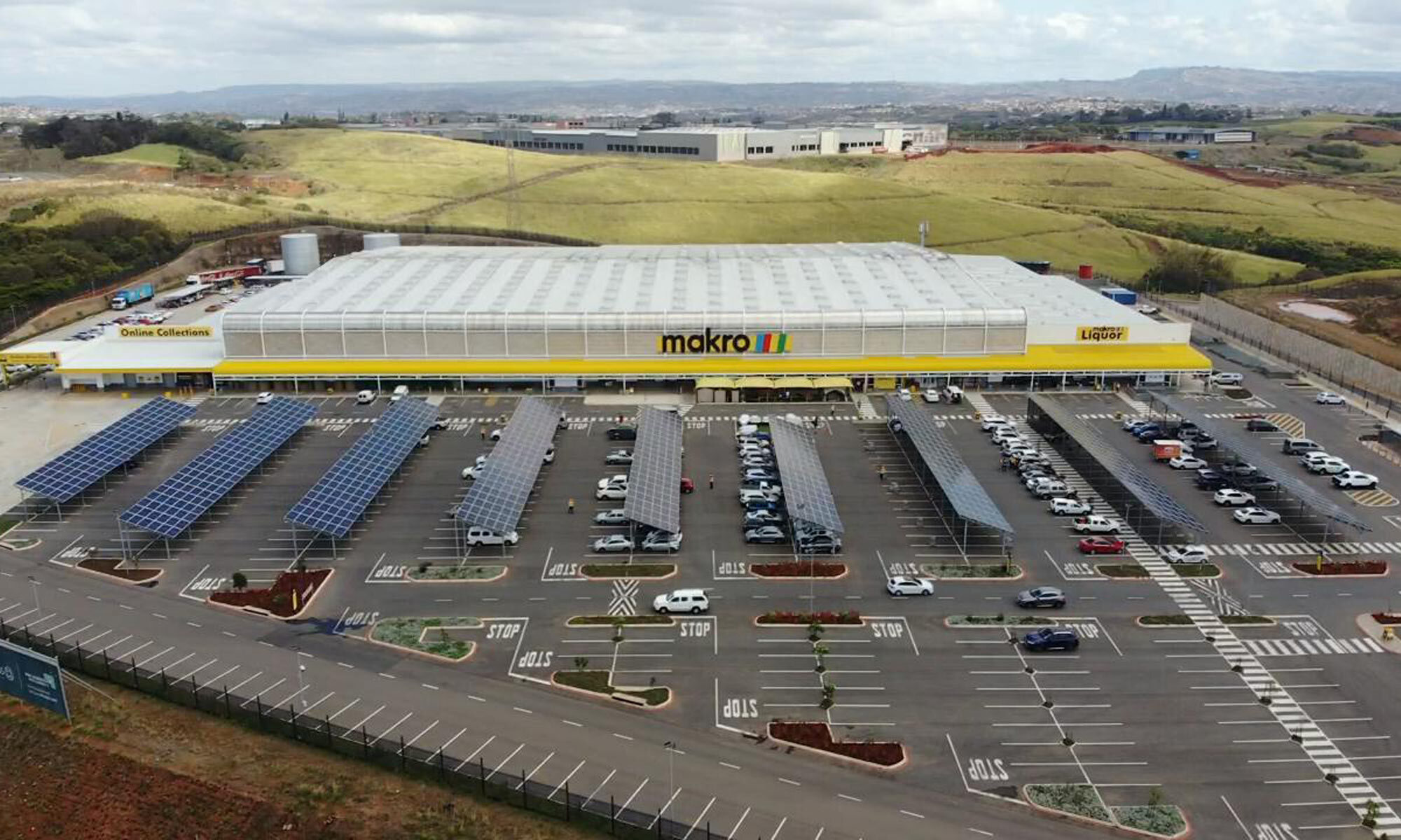 A large Makro wholesale store is shown with a spacious parking lot covered by solar panel structures. The setting is an open, semi-rural area with rolling hills in the background. Numerous cars are parked under the solar panels, and clear signage such as 'STOP' is visible on the pavement. The overall mood is utilitarian and modern, with a focus on sustainability.