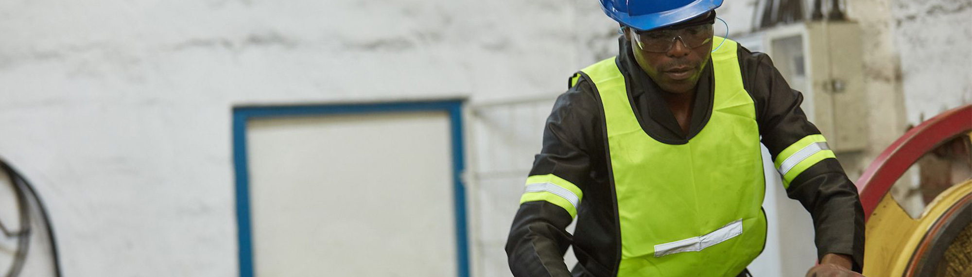 A worker wearing a bright yellow safety vest and hardhat works in a backroom.