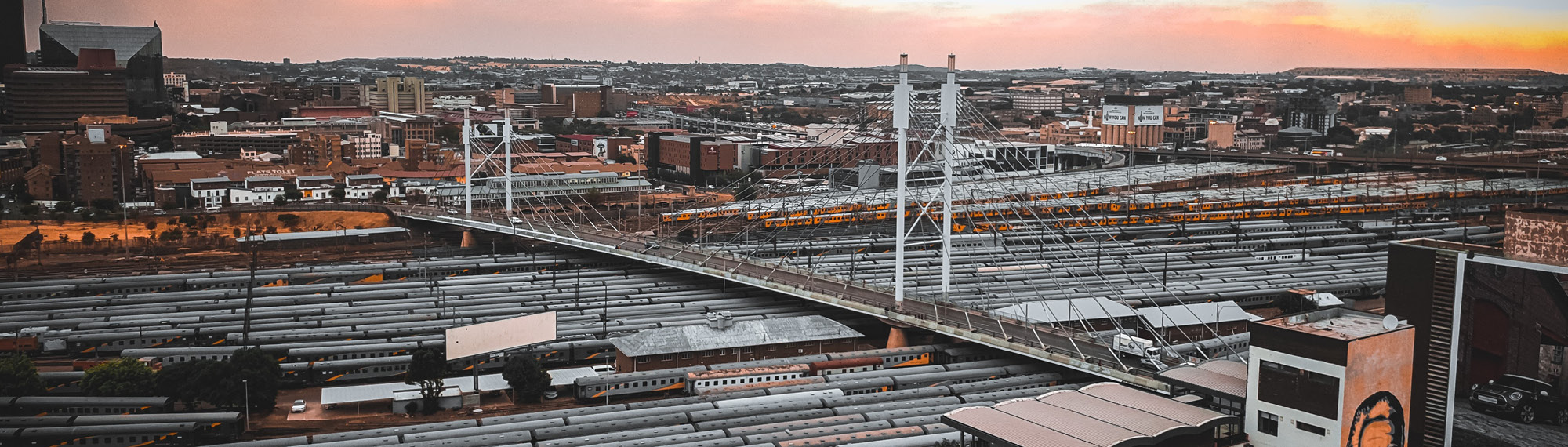 A wide shot from above, showing a bridge over a busy railyard at dusk.