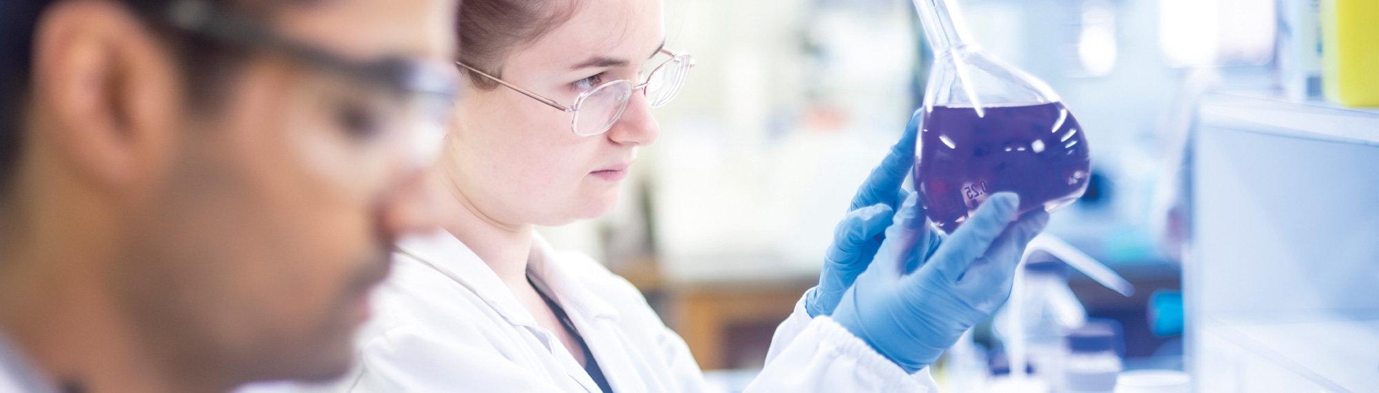 A female scientist wearing latex gloves examines a beaker full of purple liquid.